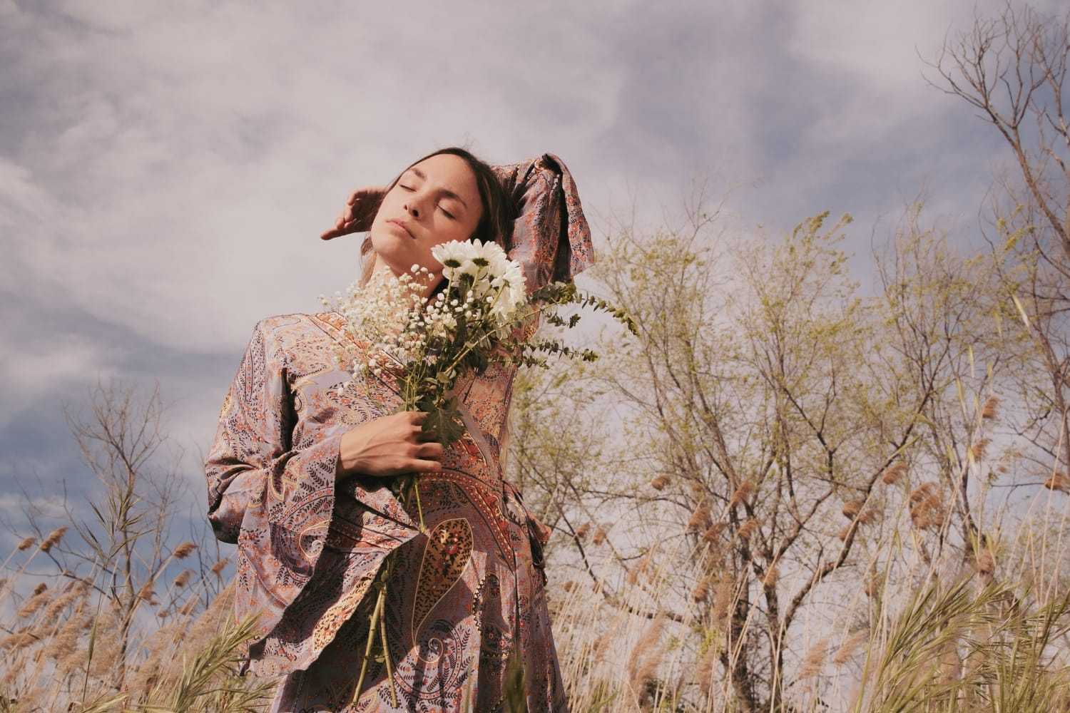 Person in patterned dress, holding wildflowers in a meadow, eyes closed under a blue sky.