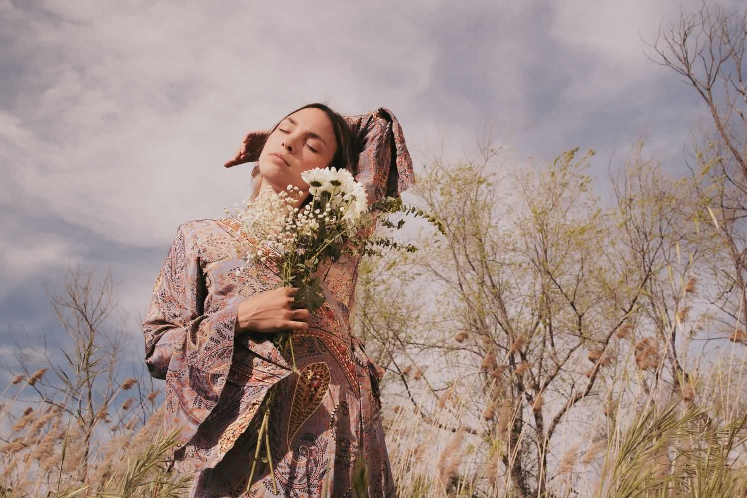 Person in patterned dress, holding wildflowers in a meadow, eyes closed under a blue sky.